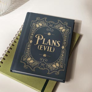 May include: A dark blue hardcover journal featuring gold foil lettering that reads PLANS (EVIL) centered within an ornate floral frame. The cover design includes small geometric shapes above and below the text, resting atop a green spiral notebook.