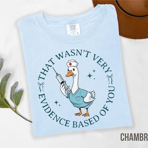 May include: Light blue Chambray t-shirt featuring a cartoon goose dressed as a nurse, holding a syringe. The text around the goose reads "THAT WASN'T VERY EVIDENCE BASED OF YOU". The shirt has a crew neck and short sleeves.