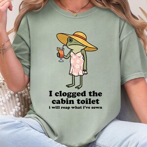May include: Sage green t-shirt featuring a cartoon frog wearing a yellow sun hat and a pink dress, holding a cocktail. Text on the shirt reads "I clogged the cabin toilet, I will reap what I've sown."