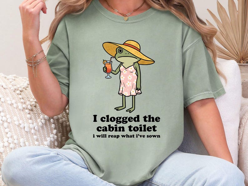 May include: Sage green t-shirt featuring a cartoon frog wearing a yellow sun hat and a pink dress, holding a cocktail. Text on the shirt reads "I clogged the cabin toilet, I will reap what I've sown."