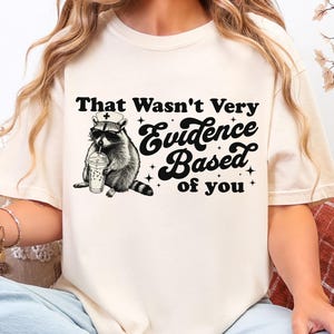 May include: Cream-colored t-shirt featuring a raccoon wearing sunglasses and a nurse's cap, holding a drink. The text "That Wasn't Very Evidence Based of you" is printed above the raccoon in black.