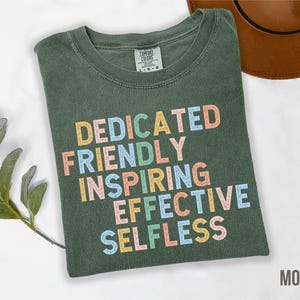 May include: A moss green t-shirt with the words "DEDICATED FRIENDLY INSPIRING EFFECTIVE SELFLESS" printed in colorful letters. The shirt is folded and lies on a white surface. A brown hat is visible in the background.
