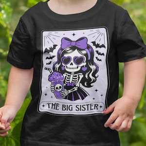 May include: Black t-shirt featuring a graphic of a skeleton girl with a purple bow, heart-shaped sunglasses, and a purple drink. The text "THE BIG SISTER" is printed below the image. Bats are in the background.