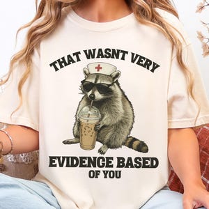 May include: Cream-colored t-shirt featuring a raccoon wearing sunglasses and a nurse's cap, holding an iced coffee. The text on the shirt reads "THAT WASNT VERY EVIDENCE BASED OF YOU."