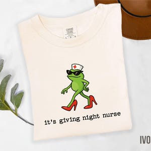 May include: Ivory-colored t-shirt featuring a cartoon frog wearing sunglasses, a nurse's cap, and red high heels. The frog is walking, and the text "it's giving night nurse" is below the frog. The shirt is made by Comfort Colors.