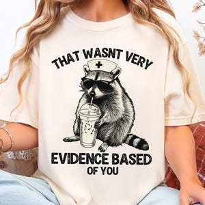 May include: Cream-colored t-shirt featuring a black and white illustration of a raccoon wearing sunglasses and a nurse's cap, holding a beverage. The text on the shirt reads "THAT WASNT VERY EVIDENCE BASED OF YOU."