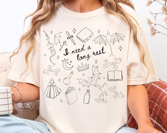 May include: Cream-colored t-shirt with a black line art design featuring dragons, books, a crown, a potion bottle, and the text "I need a long rest." The design has a fantasy theme.