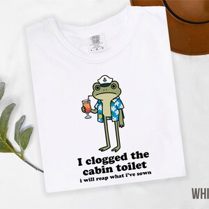 May include: White t-shirt featuring a cartoon frog wearing a captain's hat and Hawaiian shirt, holding a cocktail. Text on the shirt reads "I clogged the cabin toilet, I will reap what I've sown."