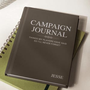 May include: A brown Campaign Journal notebook with white text that reads "CAMPAIGN JOURNAL" and "THINGS MY PLAYERS HAVE SAID SO I'LL NEVER FORGET". The name "JESSE" is printed at the bottom. A green notebook is beneath it.
