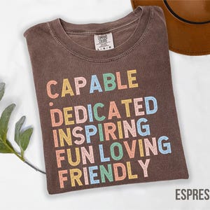 May include: Espresso-colored t-shirt with the words "CAPABLE, DEDICATED, INSPIRING, FUN, LOVING, FRIENDLY" in colorful block letters. The shirt is made of a soft material and is a comfortable fit.