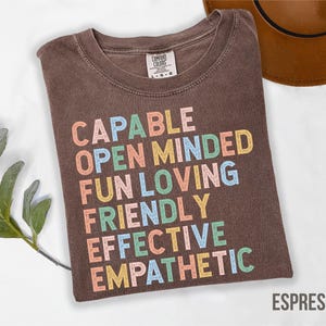 May include: Espresso-colored t-shirt with the words "CAPABLE, OPEN MINDED, FUN LOVING, FRIENDLY, EFFECTIVE, EMPATHETIC" in colorful block letters. The shirt is folded and has a tag in the upper left corner. A brown hat is in the upper right corner.
