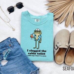May include: Seafoam green crew neck t-shirt with a cartoon frog wearing a sailor hat and Hawaiian shirt, holding a cocktail. The text "I clogged the cabin toilet, I will reap what I've sown" is printed below the frog.