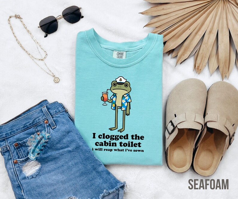 May include: Seafoam green crew neck t-shirt with a cartoon frog wearing a sailor hat and Hawaiian shirt, holding a cocktail. The text "I clogged the cabin toilet, I will reap what I've sown" is printed below the frog.