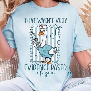 May include: Light blue t-shirt featuring a cartoon goose dressed as a nurse, holding a syringe. The goose is inside a decorative frame with the text "THAT WASN'T VERY EVIDENCE BASED of you". The shirt has short sleeves and a crew neck.