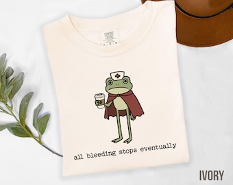 May include: Ivory-colored t-shirt featuring a cartoon frog dressed as a nurse, wearing a cape and holding a coffee cup. The text "all bleeding stops eventually" is printed below the frog. The shirt is a Comfort Colors brand.