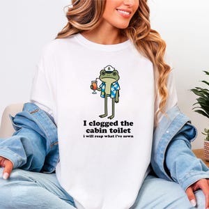 May include: White t-shirt featuring a cartoon frog wearing a sailor hat and Hawaiian shirt, holding a cocktail. The text "I clogged the cabin toilet, I will reap what I've sown" is printed below the frog. The shirt is a casual wear item.