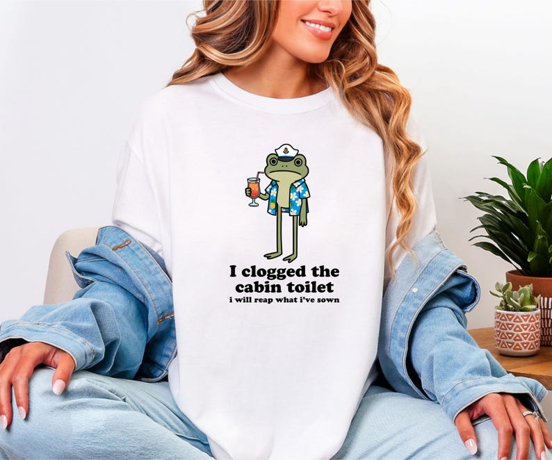 May include: White t-shirt featuring a cartoon frog wearing a sailor hat and Hawaiian shirt, holding a cocktail. The text "I clogged the cabin toilet, I will reap what I've sown" is printed below the frog. The shirt is a casual wear item.