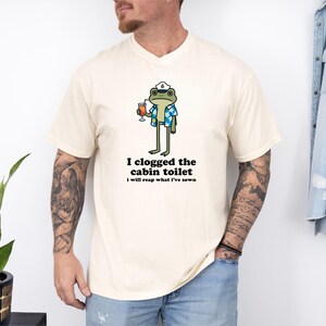 May include: Cream-colored t-shirt featuring a cartoon frog wearing a sailor hat and Hawaiian shirt, holding a cocktail. The text "I clogged the cabin toilet, I will reap what I've sown" is printed below the frog. The shirt is a casual wear item.