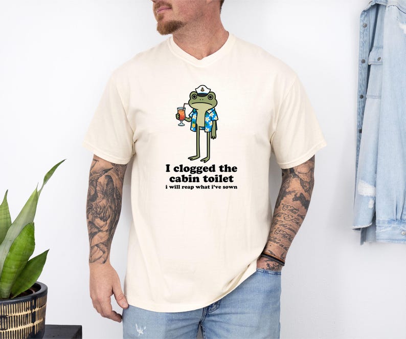 May include: Cream-colored t-shirt featuring a cartoon frog wearing a sailor hat and Hawaiian shirt, holding a cocktail. The text "I clogged the cabin toilet, I will reap what I've sown" is printed below the frog. The shirt is a casual wear item.