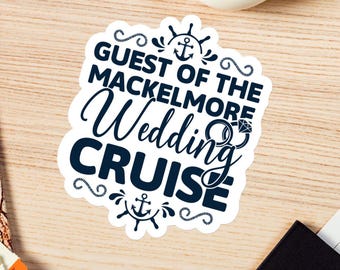 May include: A white sticker with navy blue text that reads "Guest of the Macklemore Wedding Cruise." The sticker has nautical-themed embellishments, including anchors and swirls. The sticker is on a light wood surface, with a camera and other items in the background.