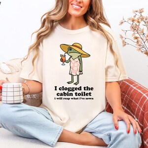 May include: Ivory-colored t-shirt featuring a cartoon frog wearing a hat and dress, holding a cocktail. The text "I clogged the cabin toilet, I will reap what I've sown" is printed below the frog. The shirt is a casual, comfortable style.
