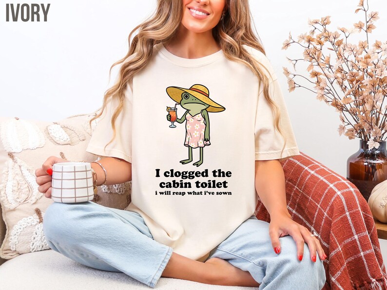 May include: Ivory-colored t-shirt featuring a cartoon frog wearing a hat and dress, holding a cocktail. The text "I clogged the cabin toilet, I will reap what I've sown" is printed below the frog. The shirt is a casual, comfortable style.