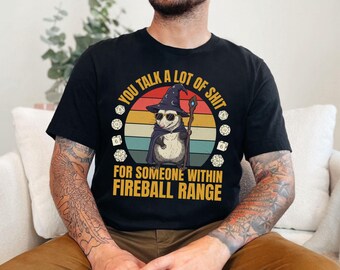 May include: Black t-shirt with a graphic of an opossum wearing a wizard hat and sunglasses, holding a staff. The text reads "YOU TALK A LOT OF SHIT FOR SOMEONE WITHIN FIREBALL RANGE". Dice are on the sides.