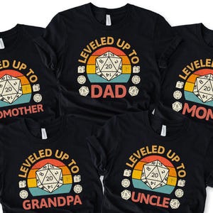 May include: Black t-shirts with a retro design featuring a D20 die and the words "Leveled Up To" followed by "Godmother", "Dad", "Mom", "Grandpa", and "Uncle". The design includes a sunset-style background.