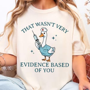 May include: Cream-colored t-shirt featuring a cartoon goose dressed as a nurse, holding a syringe. The text "THAT WASN'T VERY EVIDENCE BASED OF YOU" is arched above and below the goose illustration. The design includes small star accents.