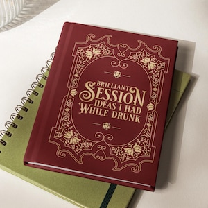 May include: A deep red hardcover journal with gold foil lettering and ornate filigree borders. The cover text reads: BRILLIANT SESSION IDEAS I HAD WHILE DRUNK, flanked by two small twenty-sided dice icons. The journal rests atop a green spiral notebook.