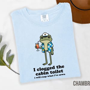 May include: Light blue t-shirt with a cartoon frog wearing a sailor hat and Hawaiian shirt, holding a cocktail. Text on the shirt reads "I clogged the cabin toilet, I will reap what I've sown." The shirt is labeled "CHAMBRAY."