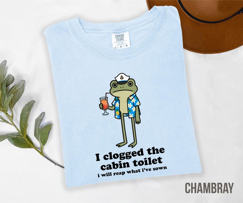 May include: Light blue t-shirt with a cartoon frog wearing a sailor hat and Hawaiian shirt, holding a cocktail. Text on the shirt reads "I clogged the cabin toilet, I will reap what I've sown." The shirt is labeled "CHAMBRAY."