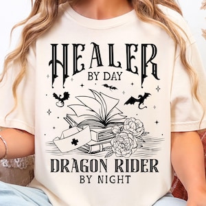 May include: Cream-colored t-shirt with a black graphic design. The design features the text "HEALER BY DAY, DRAGON RIDER BY NIGHT" with illustrations of books, bats, flowers, and a nurse's cap. The shirt is a casual, oversized fit.
