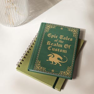 May include: A stack of notebooks with a fantasy theme. The top notebook is green with gold lettering that reads "Epic Tales of the Realm Of Custom" and a gold dragon illustration. A spiral-bound notebook and a green notebook are beneath it.