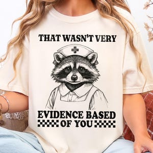 May include: Cream-colored t-shirt featuring a raccoon dressed as a nurse, wearing a nurse's cap with a cross. The text on the shirt reads "THAT WASN'T VERY EVIDENCE BASED OF YOU" in black block letters. The shirt is a casual style.