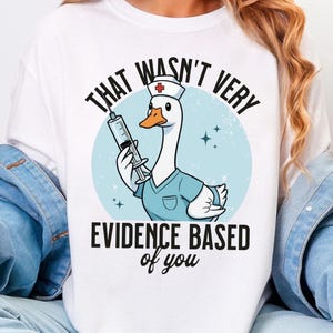 May include: White t-shirt featuring a cartoon goose dressed as a nurse, holding a syringe. The design includes the text "THAT WASN'T VERY EVIDENCE BASED of you" in black, with a light blue background.