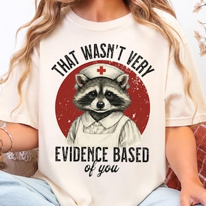 May include: Cream-colored t-shirt featuring a raccoon dressed as a nurse, set against a red circle. The text reads "THAT WASN'T VERY EVIDENCE BASED of you". The raccoon wears a nurse's cap with a red cross.