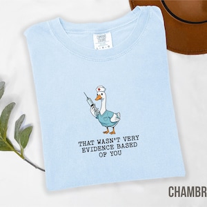 May include: Light blue t-shirt with a cartoon goose wearing a nurse's uniform and holding a syringe. The text "THAT WASN'T VERY EVIDENCE BASED OF YOU" is printed below the goose. The shirt is labeled "CHAMBRAY".