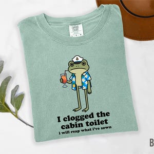 May include: A light green t-shirt with a cartoon frog wearing a captain's hat and Hawaiian shirt, holding a cocktail. The text "I clogged the cabin toilet i will reap what I've sown" is printed on the shirt.