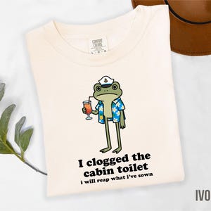May include: Ivory-colored t-shirt featuring a cartoon frog wearing a captain's hat and Hawaiian shirt, holding a cocktail. The text "I clogged the cabin toilet, I will reap what I've sown" is printed below the frog. The shirt is a Comfort Colors brand.