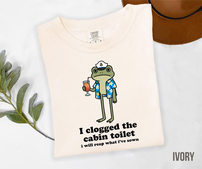 May include: Ivory-colored t-shirt featuring a cartoon frog wearing a captain's hat and Hawaiian shirt, holding a cocktail. The text "I clogged the cabin toilet, I will reap what I've sown" is printed below the frog. The shirt is a Comfort Colors brand.