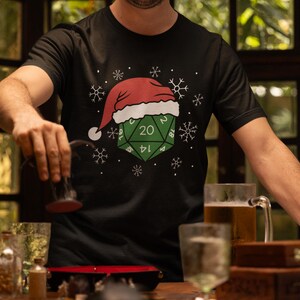 May include: Black t-shirt featuring a green D20 die with the number 20, adorned with a Santa hat and snowflakes. The shirt is a festive design, perfect for the holiday season and tabletop gaming enthusiasts.