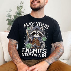 May include: Black t-shirt featuring a cartoon raccoon dressed as a fantasy character, holding a staff and surrounded by dice. The text reads "MAY YOUR ENEMIES STEP ON A D4".