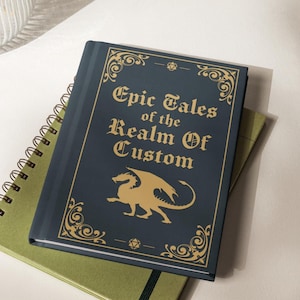 May include: A dark blue hardcover book with gold foil lettering that reads Epic Tales of the Realm Of Custom. The cover features a gold dragon illustration, ornate corner flourishes, and small geometric icons at the top and bottom center.