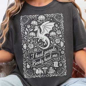 May include: Dark gray t-shirt with a white graphic featuring a dragon, flowers, and the text "I heal people. Books heal me." The design includes open books, scrolls, and a caduceus symbol, creating a whimsical and literary theme.