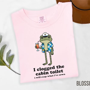 May include: A light pink t-shirt with a cartoon frog wearing a captain's hat and Hawaiian shirt, holding a cocktail. Text on the shirt reads "I clogged the cabin toilet, I will reap what I've sown." The word "BLOSSOM" is at the bottom.
