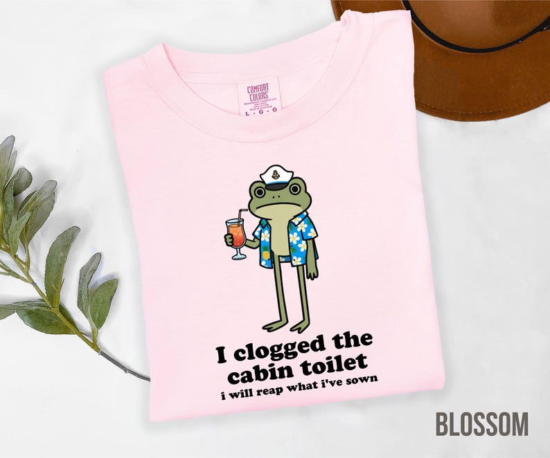 May include: A light pink t-shirt with a cartoon frog wearing a captain's hat and Hawaiian shirt, holding a cocktail. Text on the shirt reads "I clogged the cabin toilet, I will reap what I've sown." The word "BLOSSOM" is at the bottom.