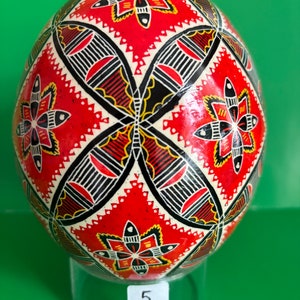 Peut inclure: Un œuf de Pâques décoré de rouge, noir et jaune avec un motif géométrique. L'œuf est sur un support en plastique transparent avec le numéro 5.