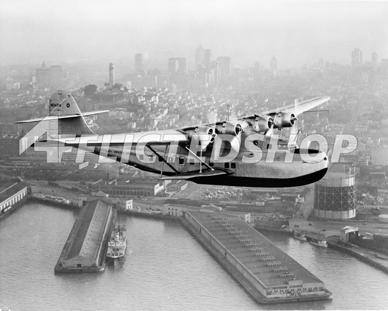 May include: A black and white photo of a Pan American World Airways Boeing 314 flying over a city skyline. The plane is in flight and the city is in the background. The plane has the words "Pan American World Airways" on the side.