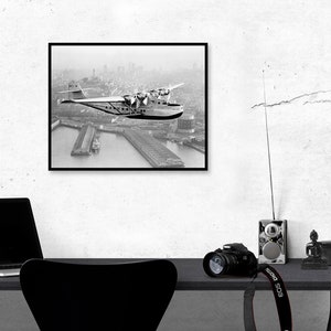 May include: Black and white photograph of a vintage seaplane flying over a city. The plane is in mid-air, with the city skyline visible in the background. The photo is framed in a black frame.